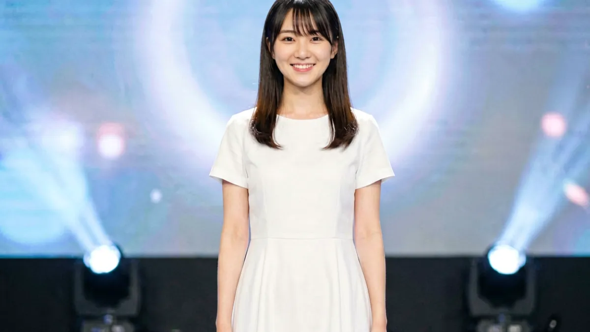 桥本香菜站在灯光明亮的舞台上微笑，身着清雅白色连衣裙，展现出她独特的清新气质